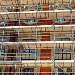 Scaffolding and temporary roofs cover the facade of a red brick building with tall windows and decorative stonework, indicating construction or renovation work in progress.