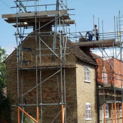 A brick house surrounded by domestic scaffolding, with two construction workers on the upper platform working on the roof under a clear blue sky. Temporary roofs protect their workspace as they complete repairs.