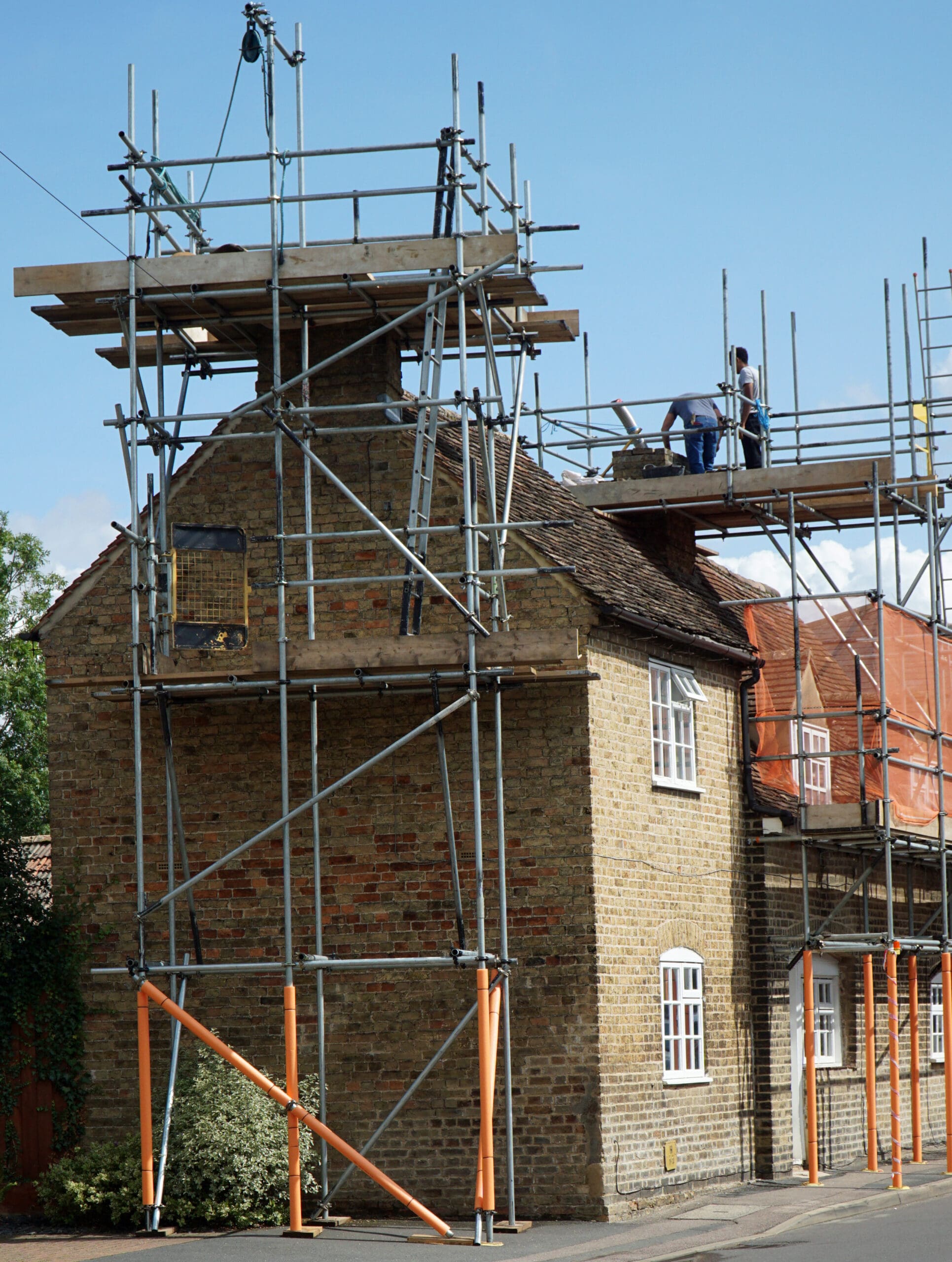 A brick house surrounded by domestic scaffolding, with two construction workers on the upper platform working on the roof under a clear blue sky. Temporary roofs protect their workspace as they complete repairs.