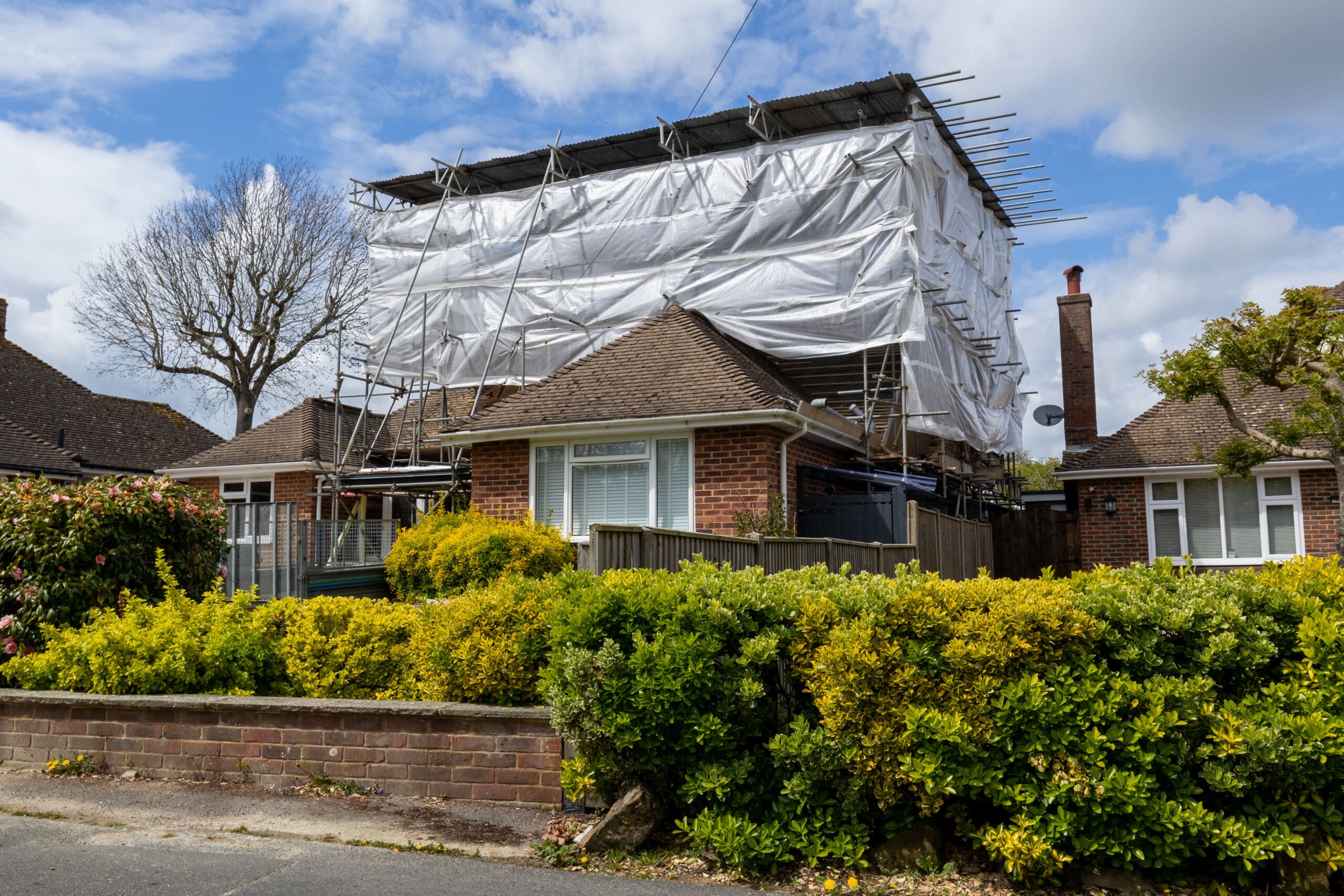 A brick house with a tiled roof is covered in commercial scaffolding and white sheeting, likely for renovation or roof work. The front yard is lined with green bushes, and temporary roofs provide added protection while work is underway.
