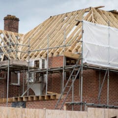 A brick house under construction, surrounded by domestic scaffolding and ladders. The roof is partially built with visible wooden beams, some exterior is covered with white sheeting, and a chimney wraps installation is underway.