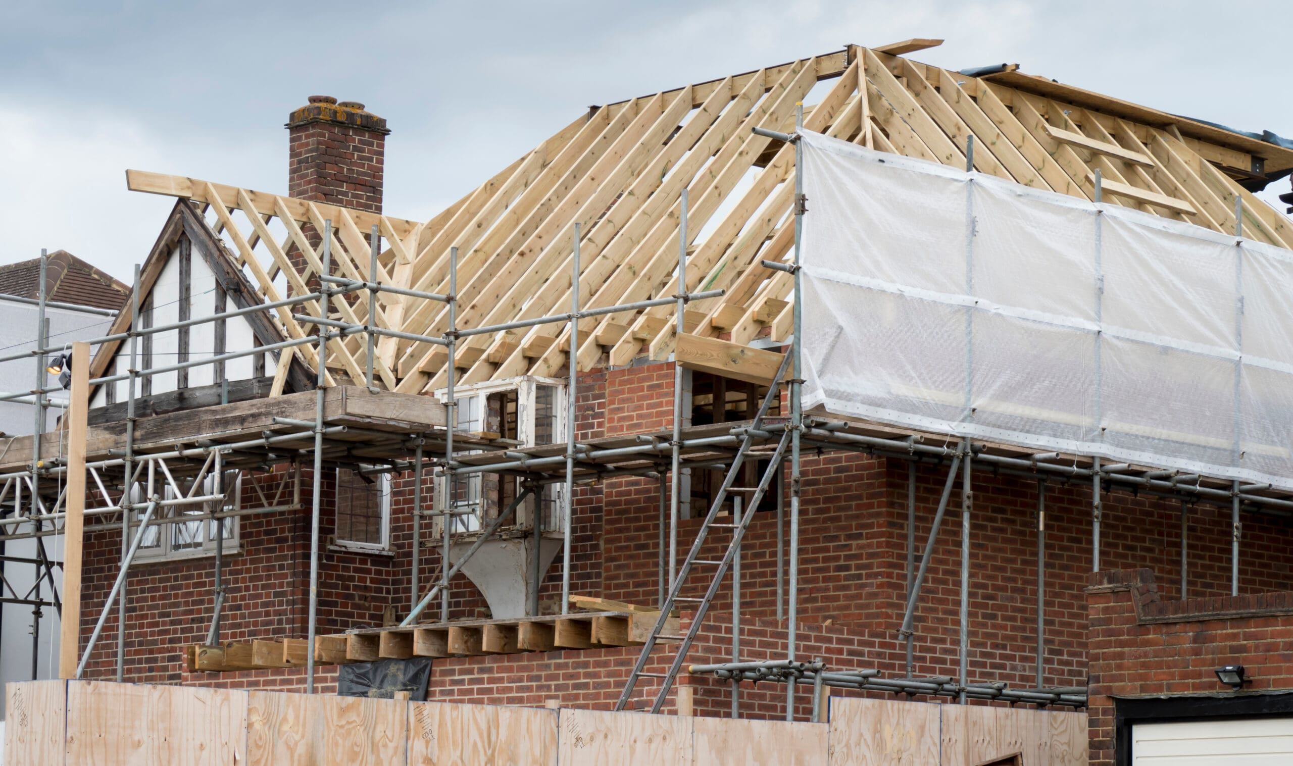 A brick house under construction, surrounded by domestic scaffolding and ladders. The roof is partially built with visible wooden beams, some exterior is covered with white sheeting, and a chimney wraps installation is underway.