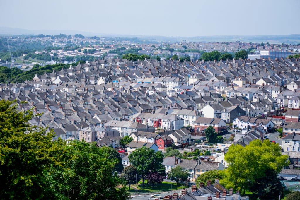 A densely packed residential neighborhood with rows of grey-roofed houses, some featuring temporary roofs and domestic scaffolding, interspersed with trees and greenery, stretching into the distance under a hazy sky.