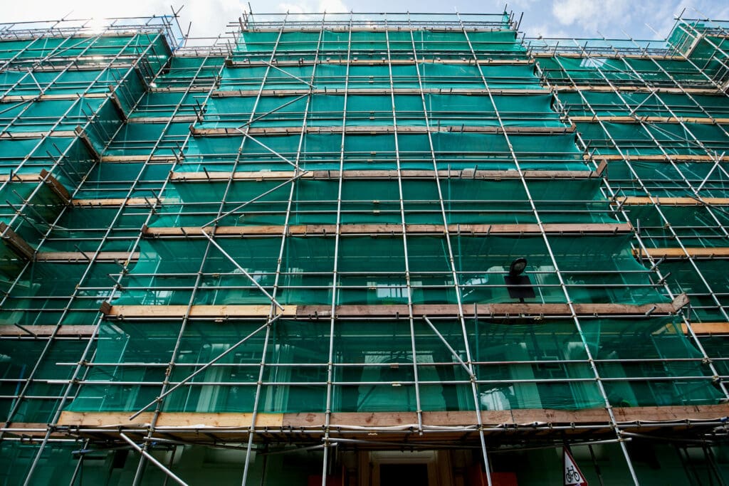 A building facade covered in green safety netting and surrounded by multiple levels of commercial scaffolding, seen from below against a partly cloudy blue sky.
