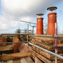 A brick chimney stack with two red chimney pots, both covered with wire guards, is surrounded by metal scaffolding on a rooftop overlooking houses and trees under a partly cloudy sky.