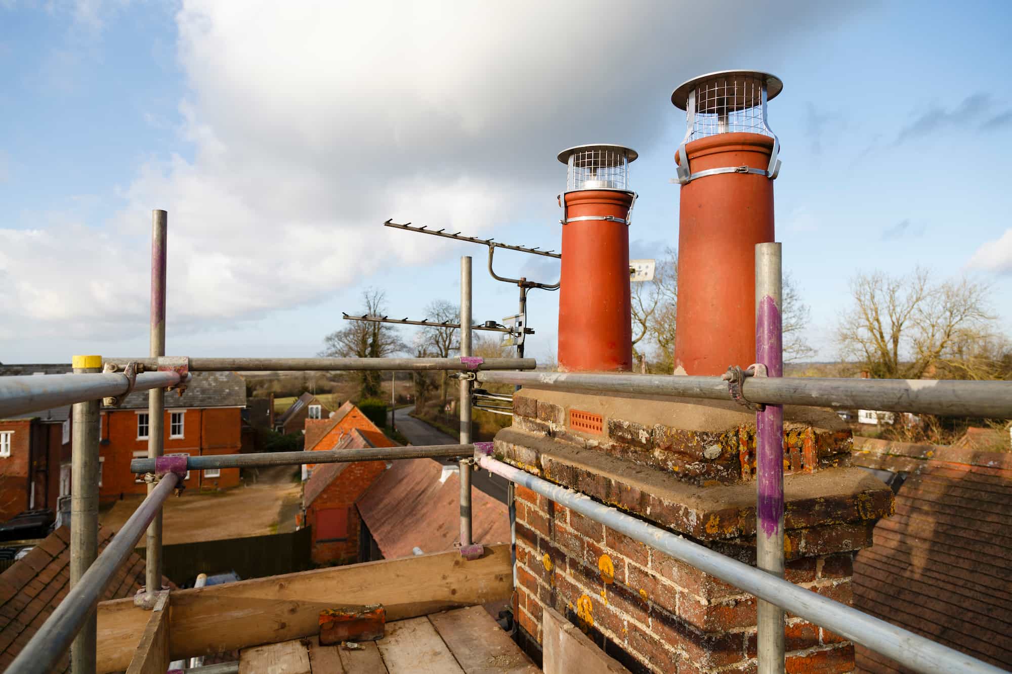 A brick chimney stack with two red chimney pots, both covered with wire guards, is surrounded by metal scaffolding on a rooftop overlooking houses and trees under a partly cloudy sky.