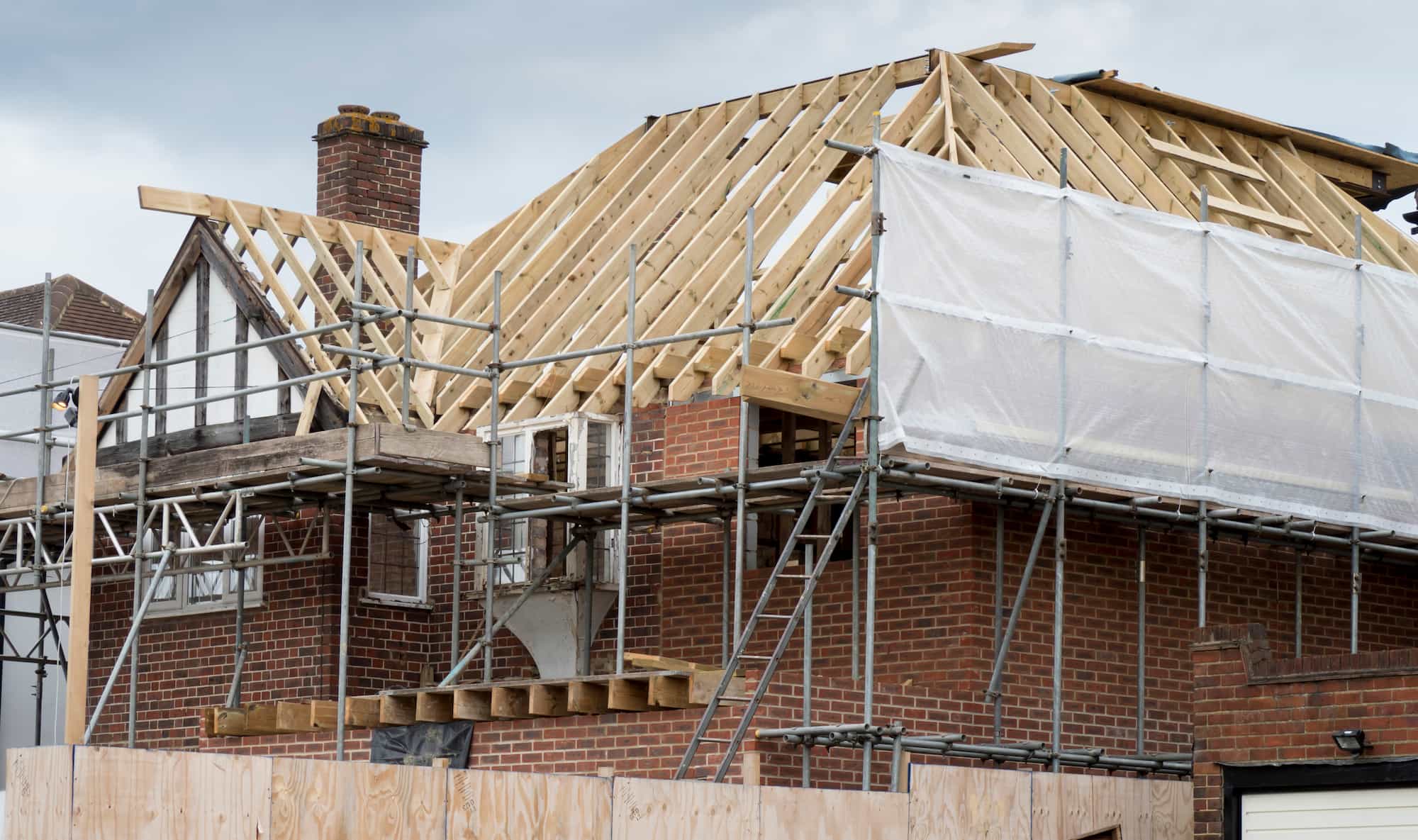 A brick house under renovation, with scaffolding and exposed wooden roof beams. Part of the roof is covered with plastic sheeting, and construction materials are visible around the building.