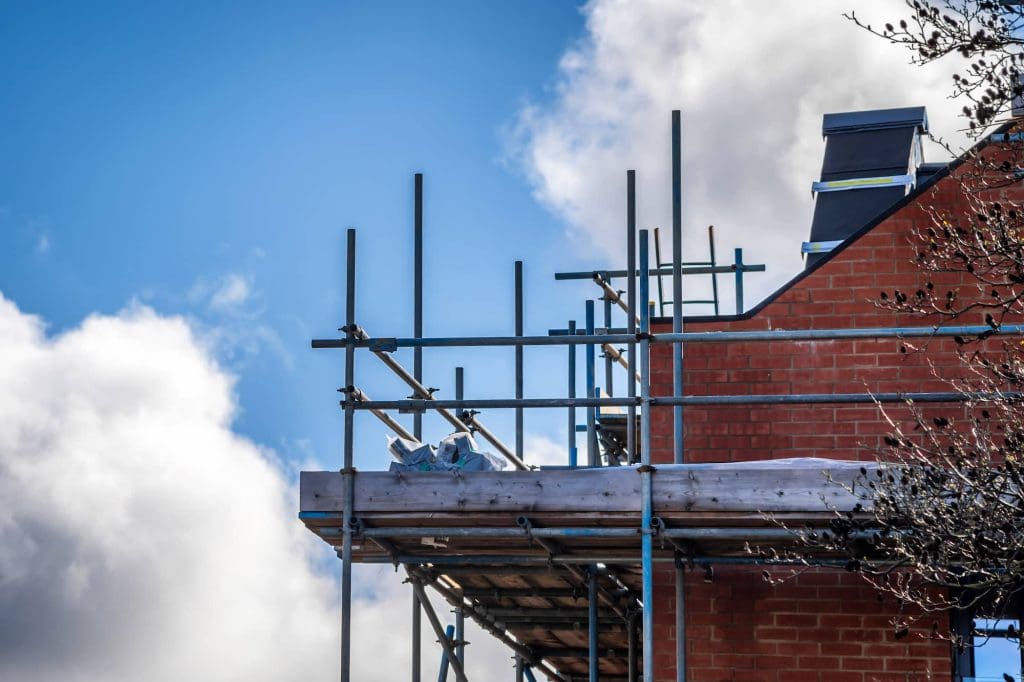 Scaffolding surrounds the upper floor of a brick building under construction, with a blue sky and white clouds in the background.