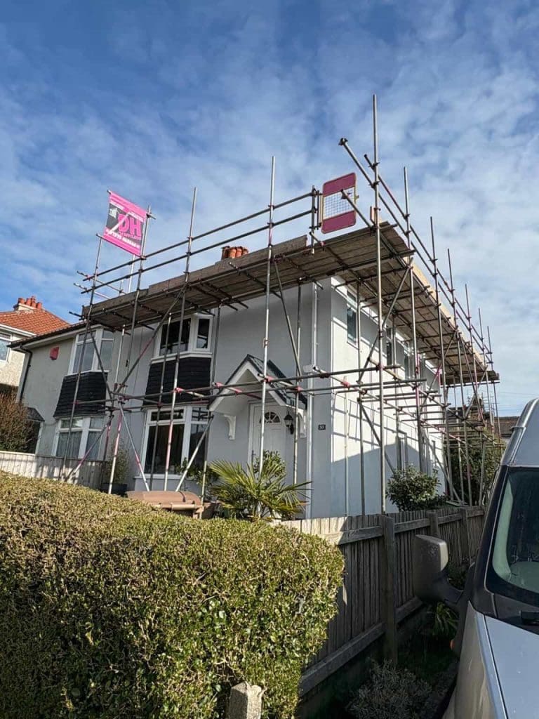 A two-story white house surrounded by scaffolding with pink company signs on top, indicating construction or renovation work. The house is behind a hedge with a blue sky and some clouds in the background.