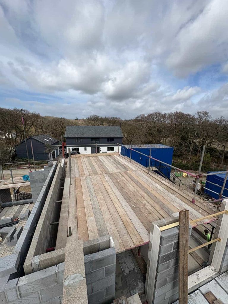 A construction site with unfinished cinder block walls and a wooden plank floor. In the background, there's a white house with a dark roof, blue containers, trees, and a cloudy sky overhead.