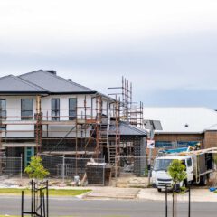 A two-story house under construction with scaffolding, a truck parked in front, construction materials scattered around, and nearby completed homes.