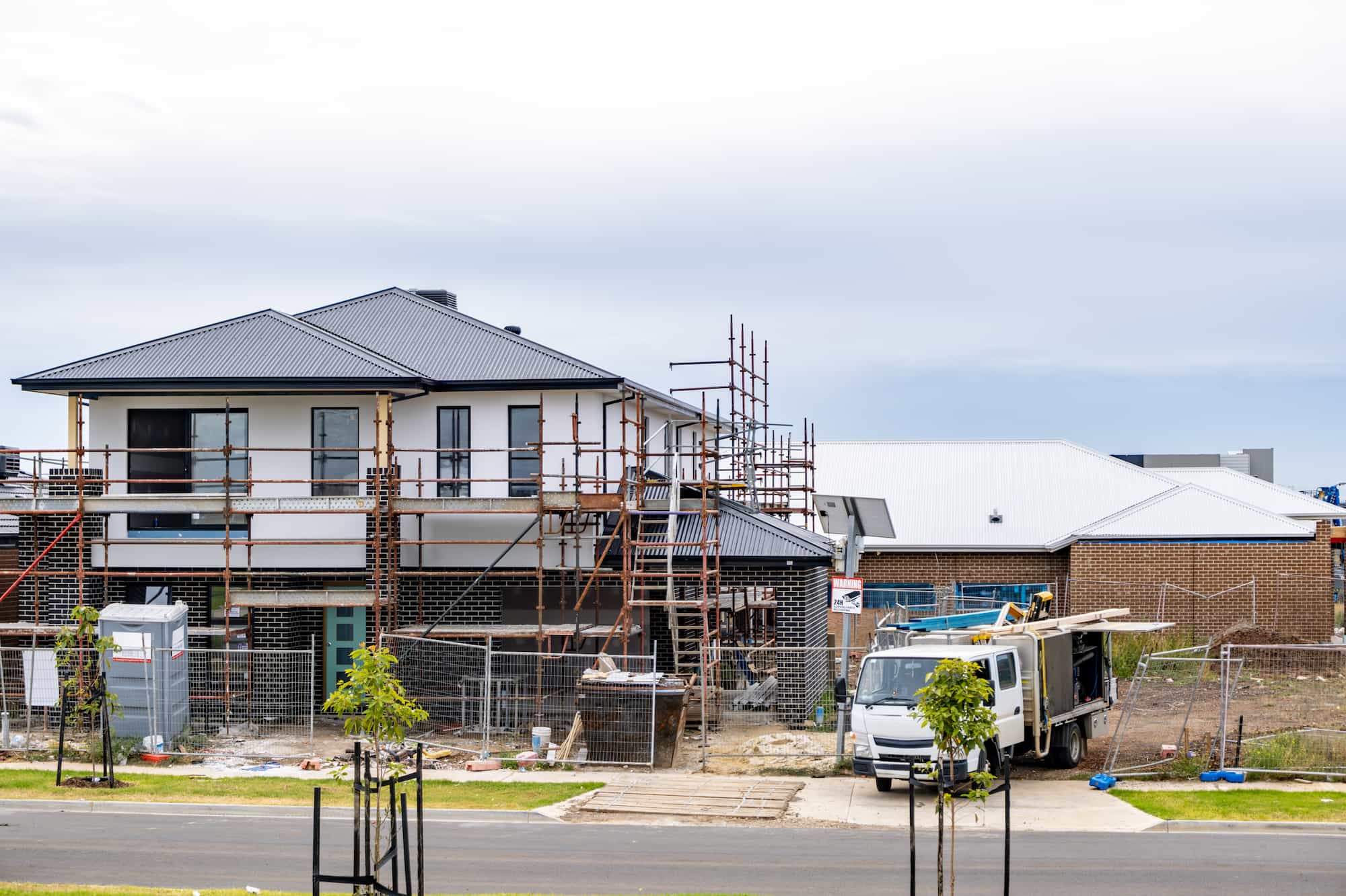 A two-story house under construction with scaffolding, a truck parked in front, construction materials scattered around, and nearby completed homes.