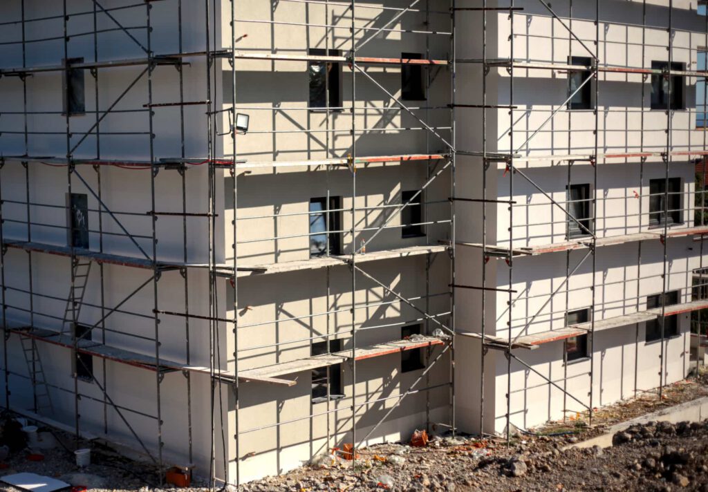 A multi-story building under construction with scaffolding around the exterior. The ground below is covered with debris and construction materials.