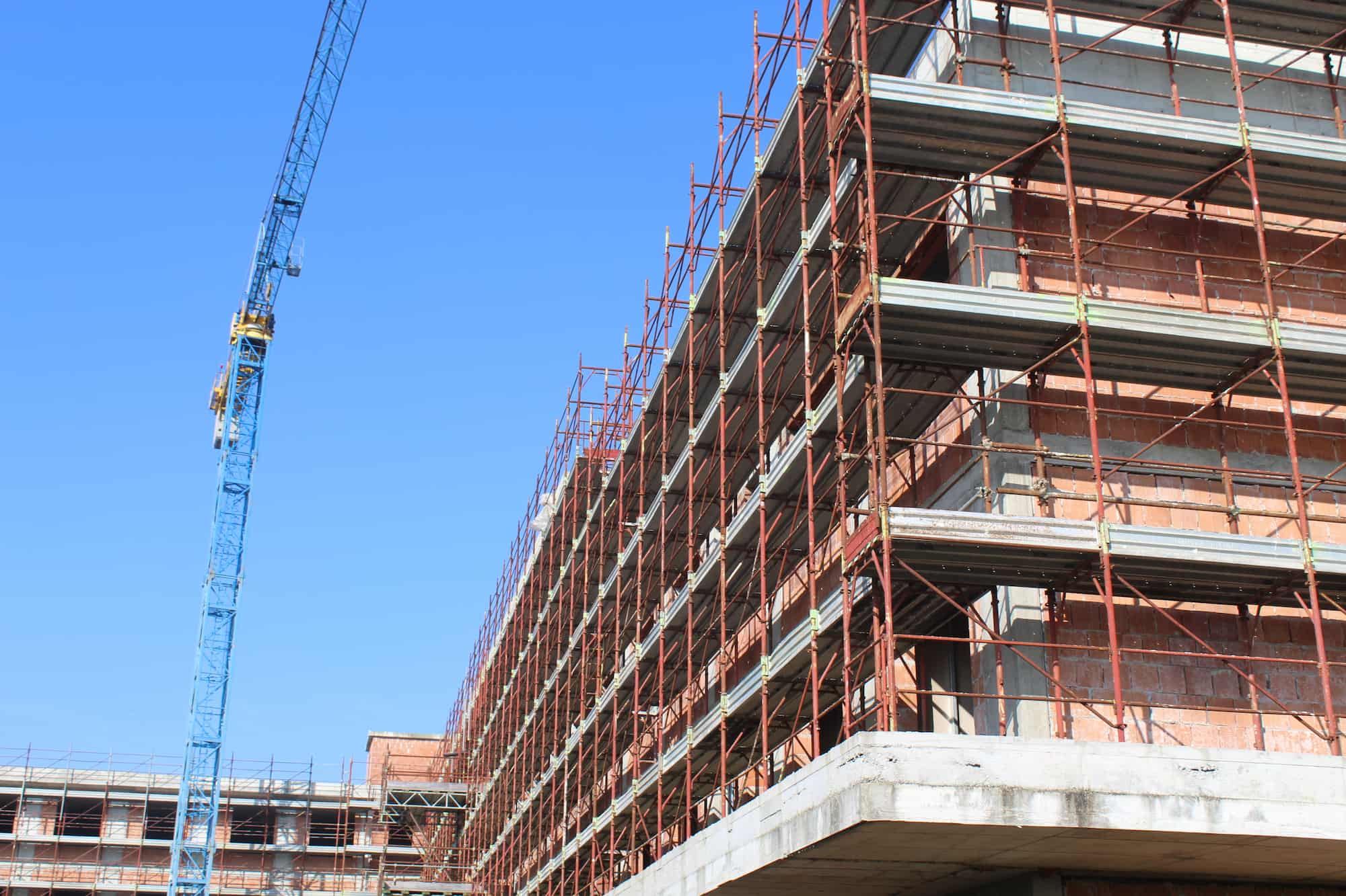 A large building under construction with red scaffolding and a blue crane against a clear blue sky. The structure appears to be made of concrete and brick.