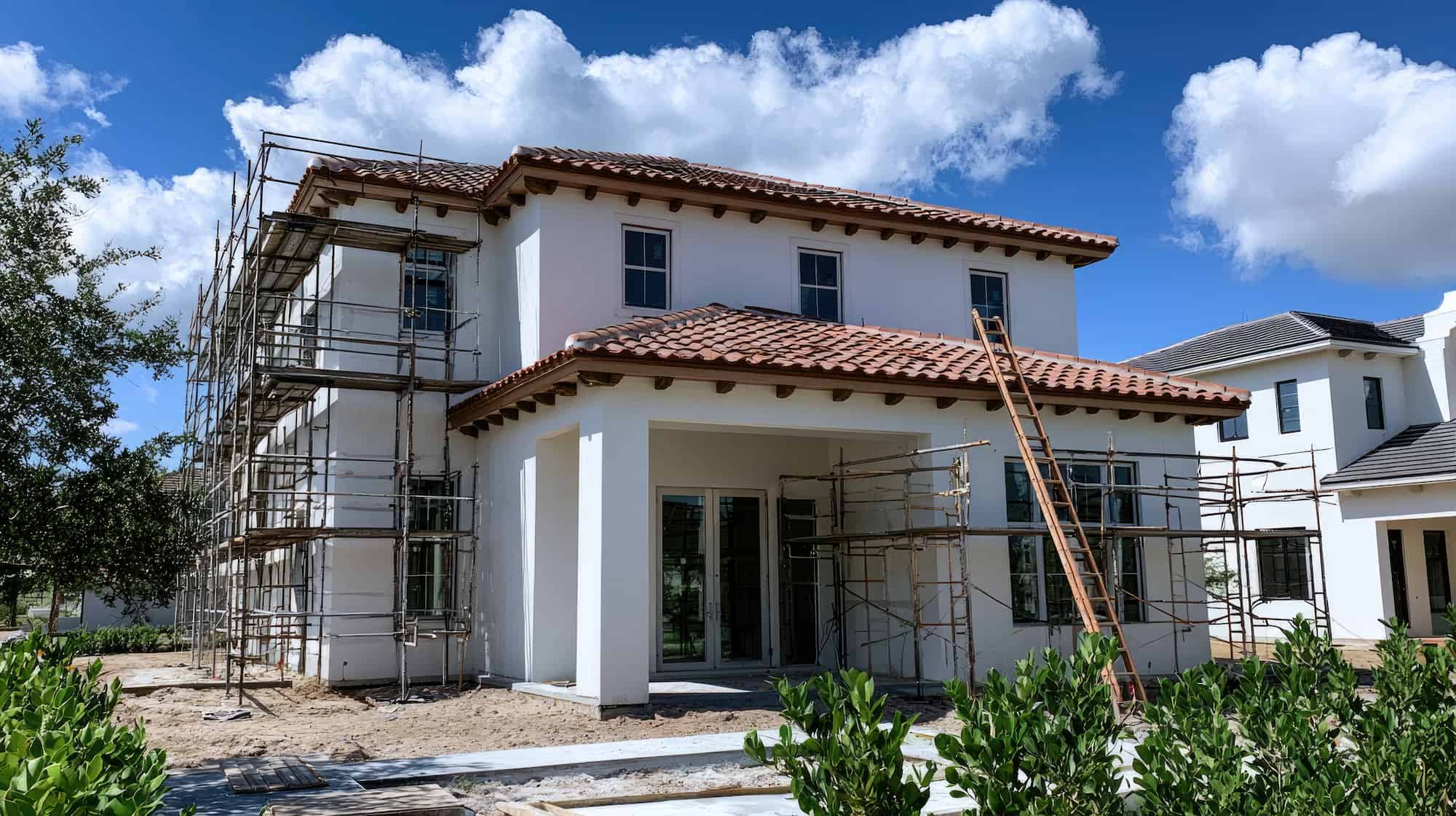 Two-story house under construction with scaffolding on multiple sides, red tile roof, white exterior walls, and a clear blue sky with clouds in the background. Shrubs are in the foreground.