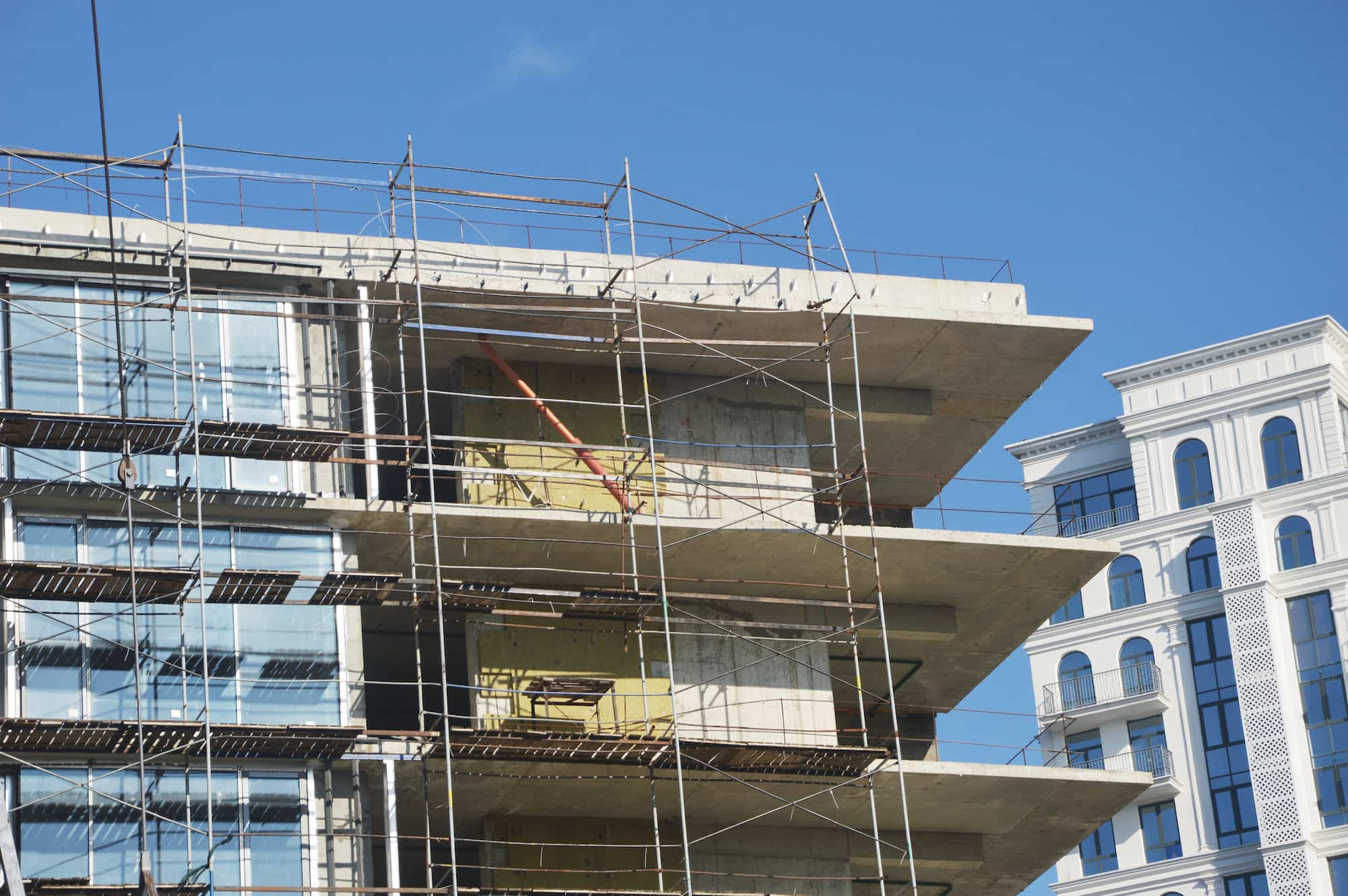 A modern building under construction with scaffolding on the exterior, next to a finished white building with many windows, against a clear blue sky.