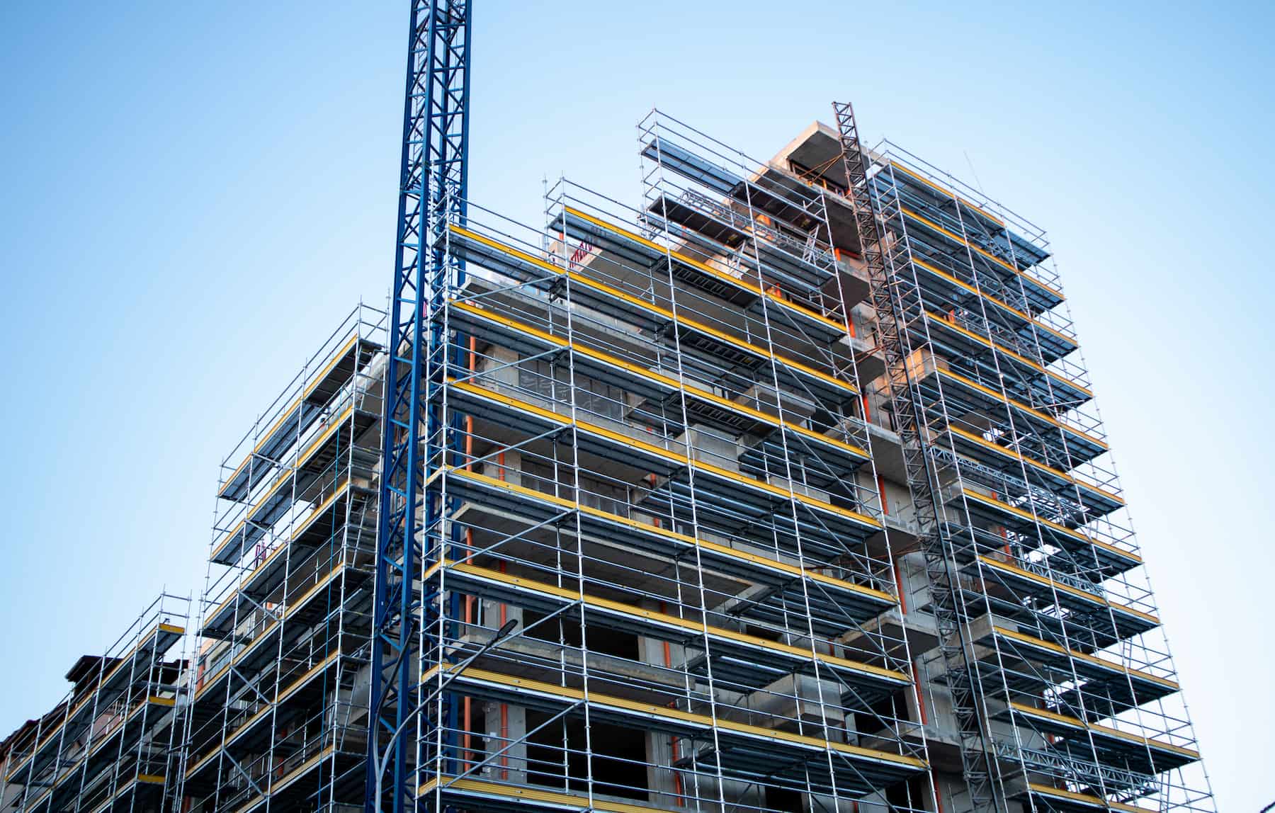 A multi-story building under construction with visible scaffolding and a blue crane beside it, set against a clear blue sky.