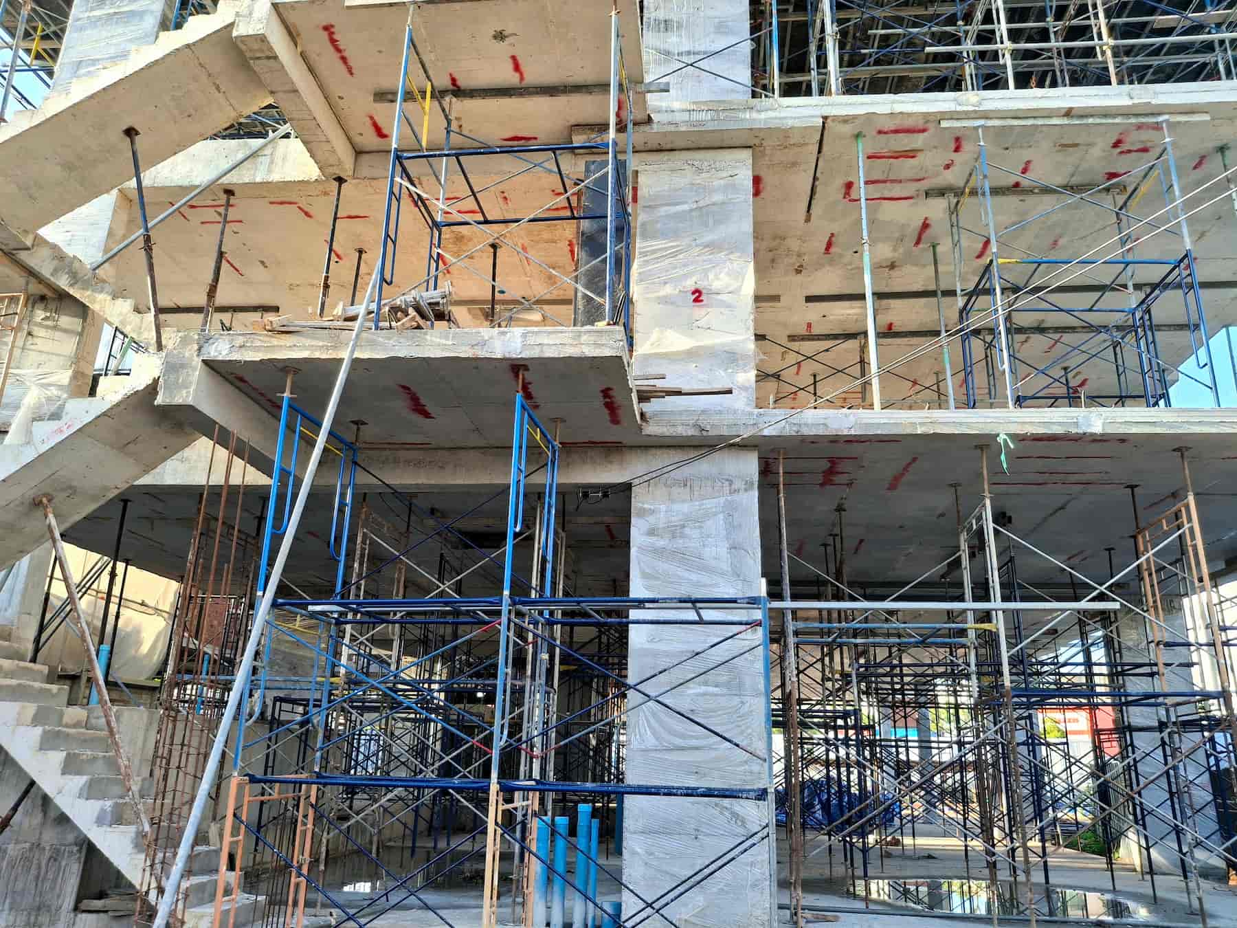 Concrete building under construction with exposed floors, pillars, staircases, and scaffolding. Blue, black, and orange metal supports are visible, along with markings on the concrete. No workers are shown.