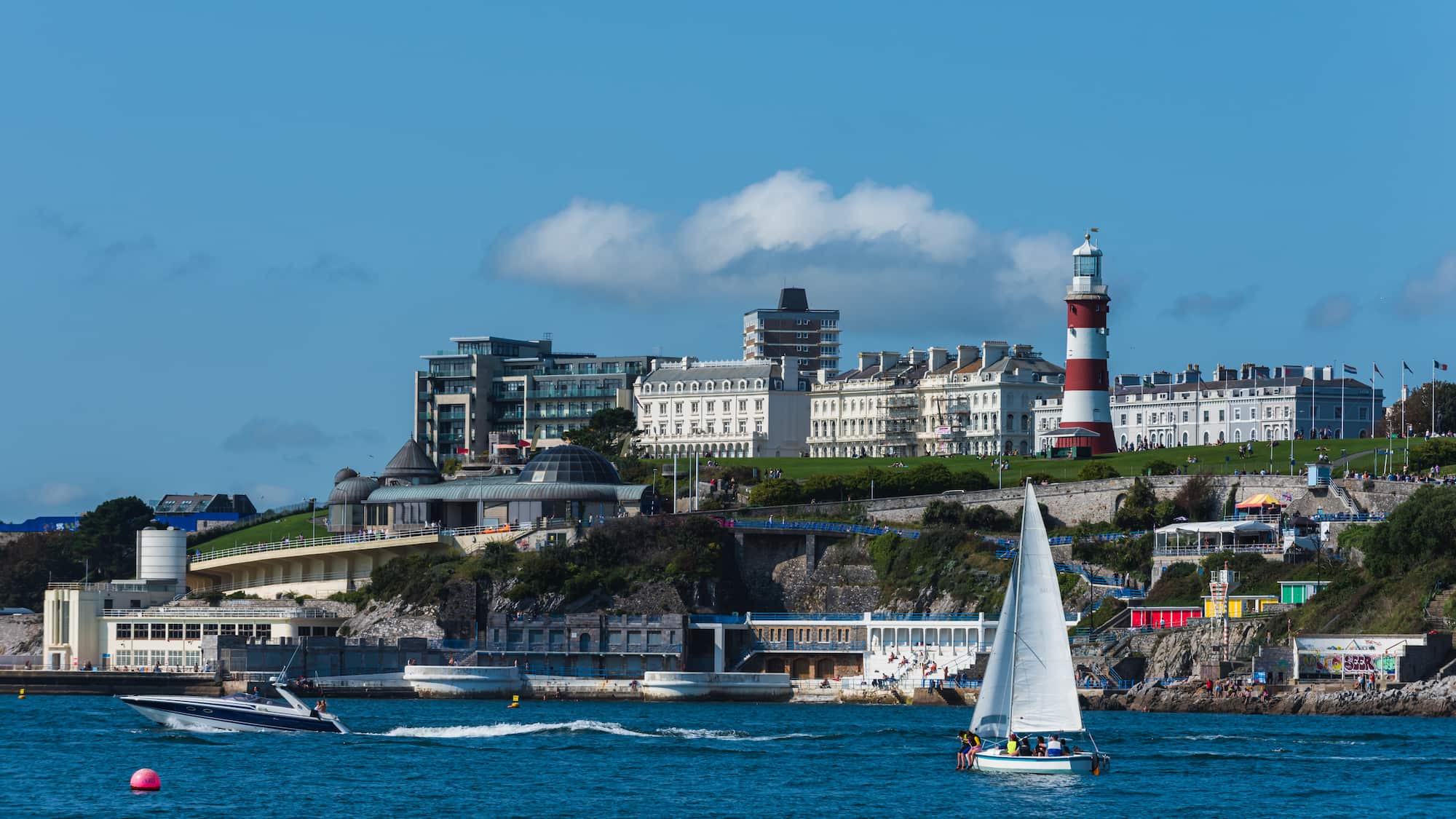 A sailboat and a motorboat on blue water with a seaside town, white buildings, and a red and white lighthouse on a hill under a blue sky with scattered clouds.