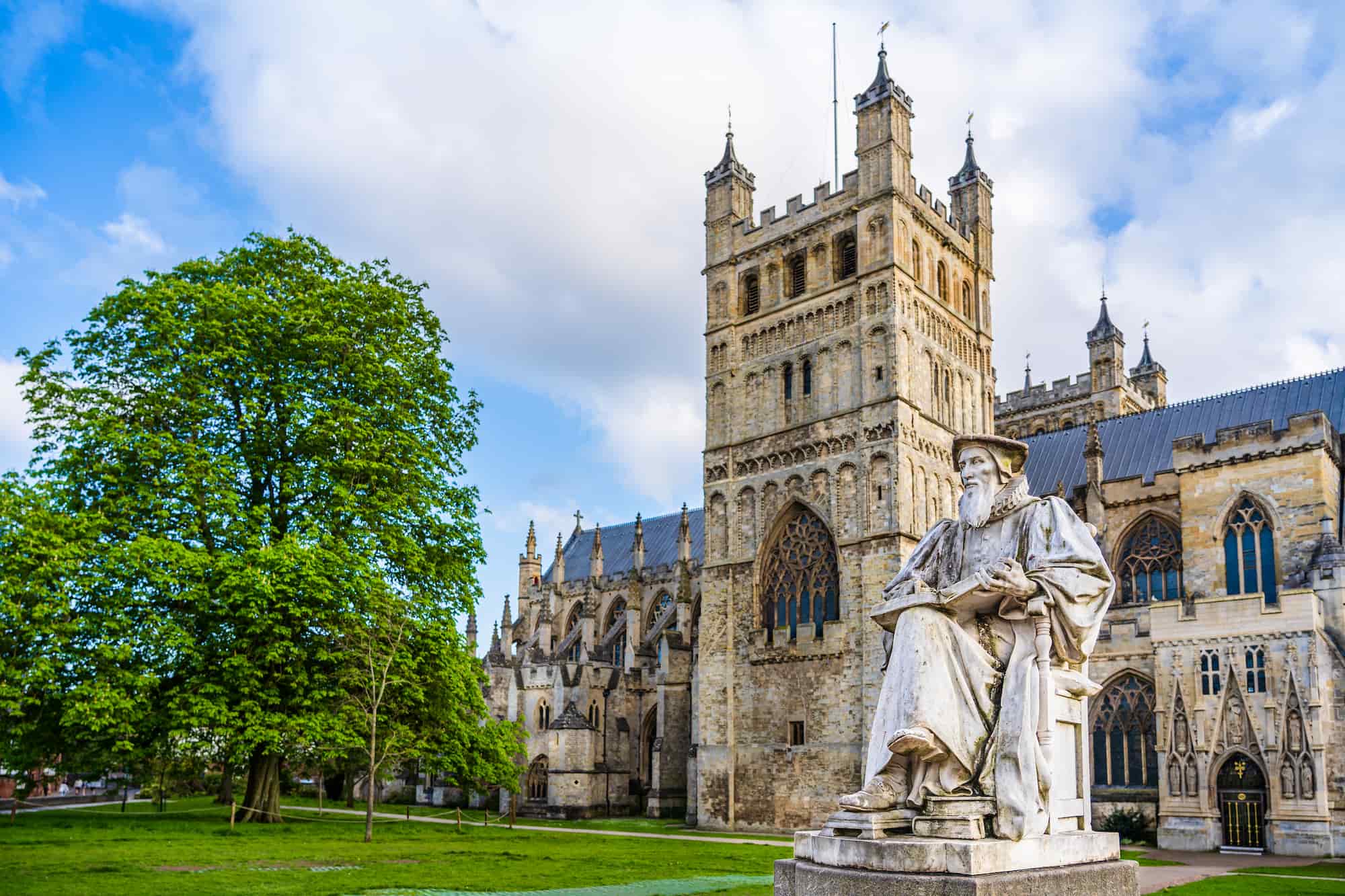 A large, ornate cathedral with tall towers stands beside a leafy green tree; a stone statue of a seated figure is in the foreground under a blue sky with clouds.