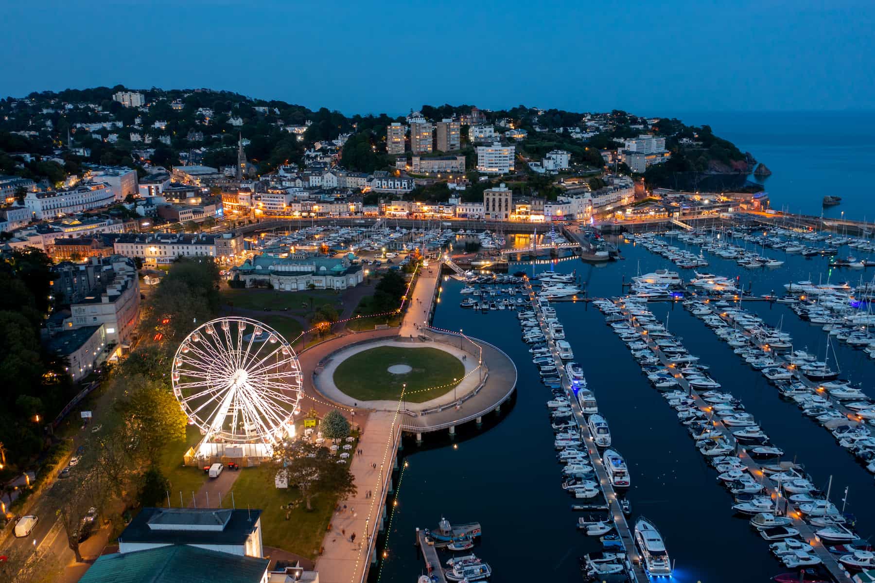 Aerial view of a marina at dusk with many docked boats, a lit Ferris wheel, and a coastal town with illuminated buildings on hills by the sea.
